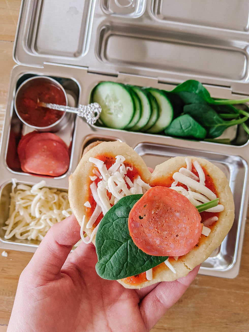 A planetbox brand lunch box is shown, a stainless stell lunch box with various sized compartments to hold different lunch items. The box is filled with marinara sauce in a small dish with a small spoon, pepperoni, spinach, shredded cheese, and some cucumbers. A heart-shaped pizza is being held up above the lunch box, and it is decorated with the marinara sauce, shredded cheese, a piece of spinach, and a piece of pepperoni
