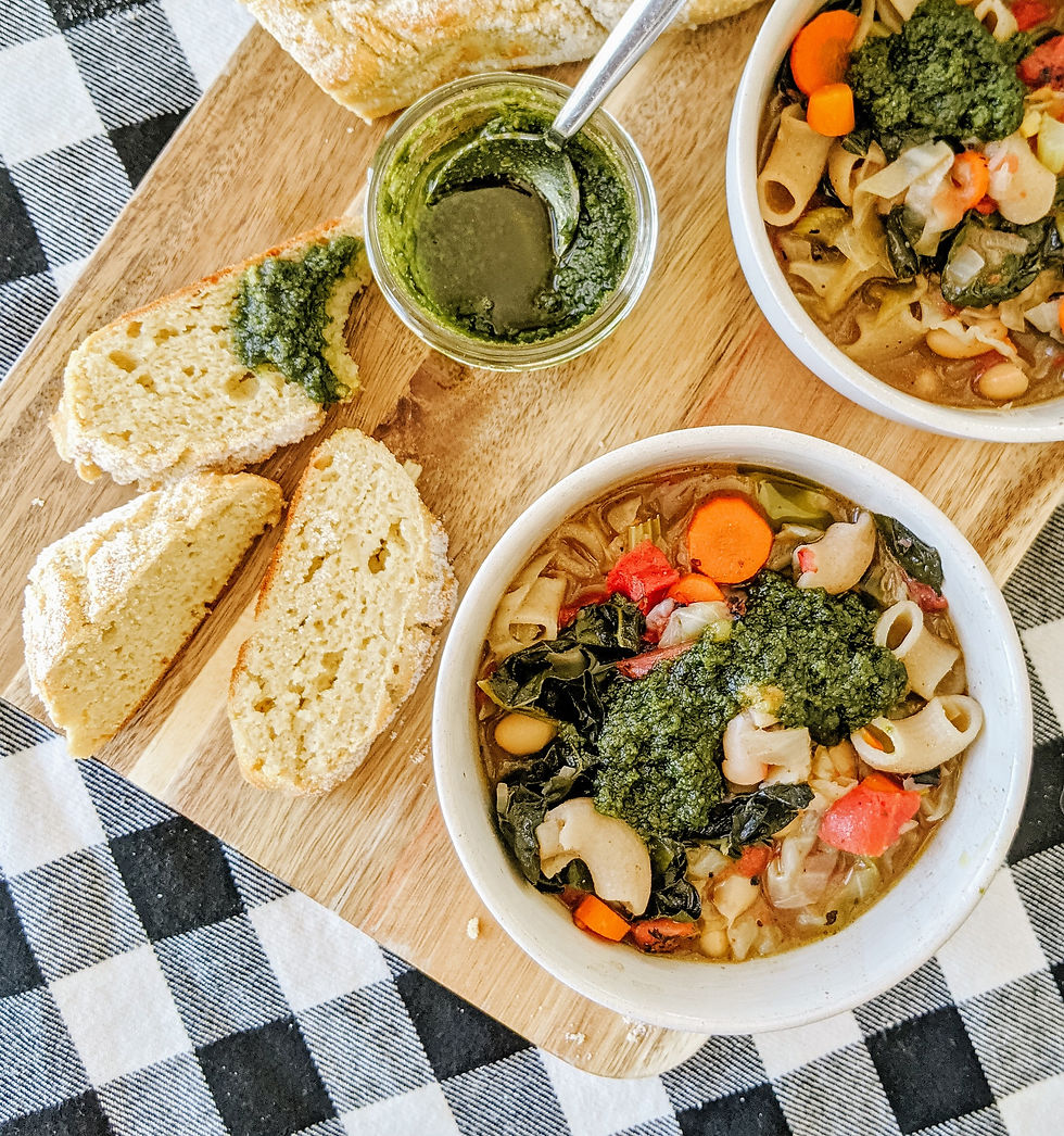 a close up of one bowl. Two bowls of minestrone soup are sitting on top of a wooden cutting board slab. The soup has elbow style pasta inside, bright red tomatoes, and carrots. Each bowl is topped with a dollop of green pesto. There's also a cassava flour baguette to the side, partly sliced, with pesto dipped. The background is a white and black buffalo check table cloth