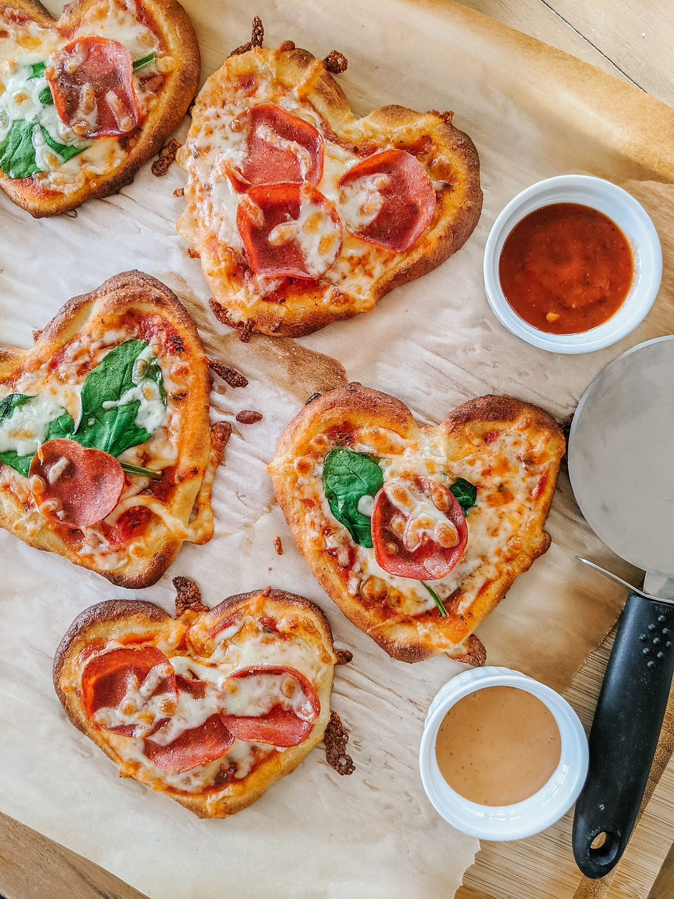 Rows of mini, heart-shaped pizzas are shown in an overhead view. All pizzas have a combination of pepperoni, warm bubbly cheese, and spinach. There's two dips in small containers, one is orange and the other is red. There's a pizza wheel off to the side.