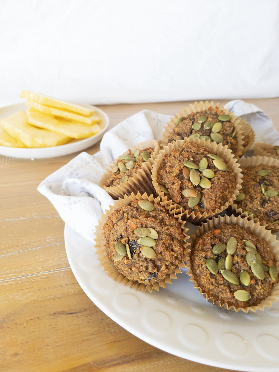 A close up of a platter of grain-free, dairy-free, gluten-free, refined sugar free, and paleo Morning Glory Muffins. You can see a plate of pineapple in the background. The muffins are topped with pepitas