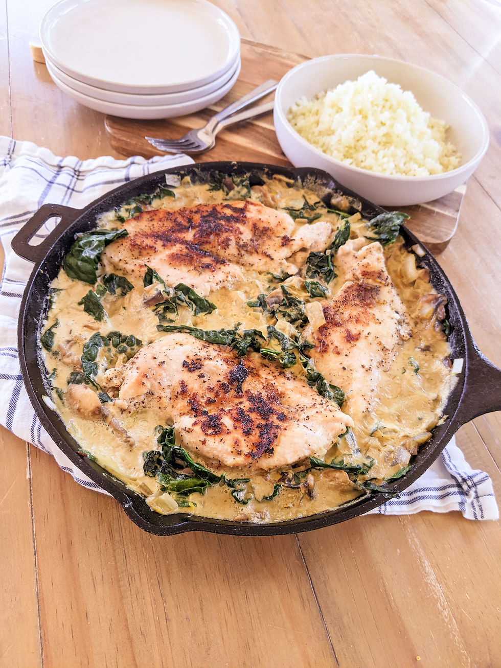 A cast iron pan is shown with three seared chicken breasts simmered in a creamy dairy-free mushroom sauce with kale. There's a bowl of cauliflower rice to serve it over.