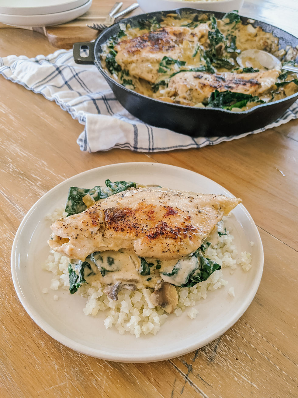 a plate of cauliflower rice with a dairy free, creamy mushroom and kale sauce and a seared chicken breast on top is shown. In the background is the rest of the meat in a cast iron skillet.