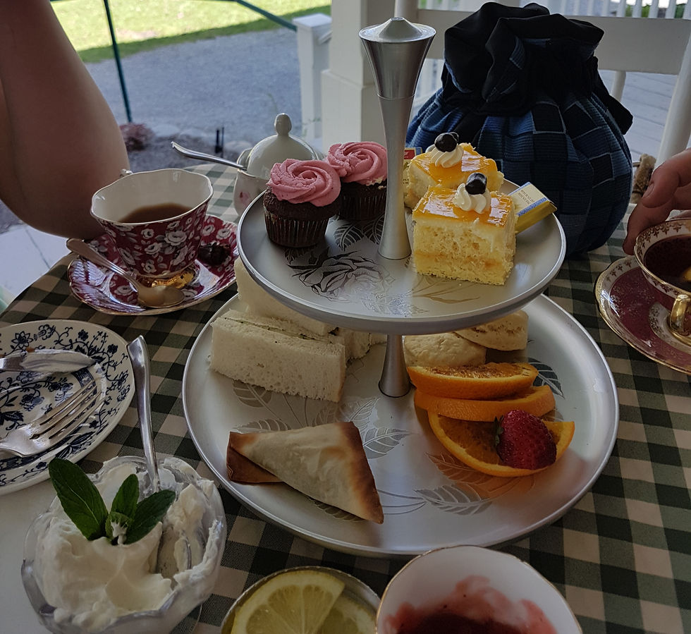 A tiered tray with cupcakes, sandwiches, and fruit slices on a checkered tablecloth. Two floral teacups with tea beside it. Outdoor setting.