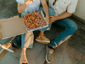 Romantic rooftop couples session with casual outfits and pizza picnic. A couple sits close together on the concrete, sharing a pepperoni pizza straight from the box during their engagement photo shoot. Casual summer date night vibes with golden hour light, laid-back outfits, and candid photography style — perfect inspiration for cozy couples photoshoot ideas.