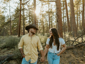 Engaged couple holding hands and smiling at each other in a sunlit pine forest in Oregon during their rustic outdoor engagement session. The man wears a cowboy hat and yellow shirt, while the woman wears a white blouse and light jeans, surrounded by tall trees and golden evening light.
