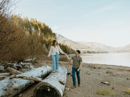 Adventurous couple walking across large driftwood logs by the water’s edge, surrounded by Oregon mountain scenery