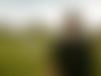 A candid engagement photo of a woman in a white dress playfully twirling in a green meadow while her partner watches from the foreground, set against an overcast sky and tree-lined Oregon landscape.