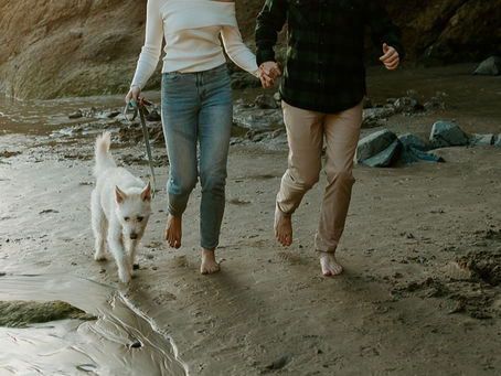 Barefoot couple walking hand-in-hand with their white terrier on an Oregon coast beach at low tide, golden hour light and coastal rocks in the background.