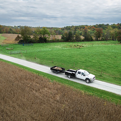 BWise Dump Trailer full of logs being pulled down a country road next to a corn field.
