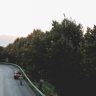 En slingrande väg med en bil och en vespa. Vägen är kantad av grönska och stenhus skymtas i bakgrunden. Umbrien italien.