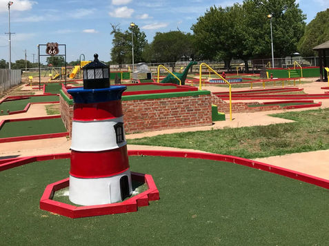 A colorful 18-hole miniature golf course at Sayre City Park under a bright blue sky. In the foreground, a red, white, and blue lighthouse obstacle sits on a green artificial turf green. In the background, other classic obstacles are visible, including a green dinosaur and a brown Route 66 highway sign. The course is surrounded by red brick borders and lush green trees.