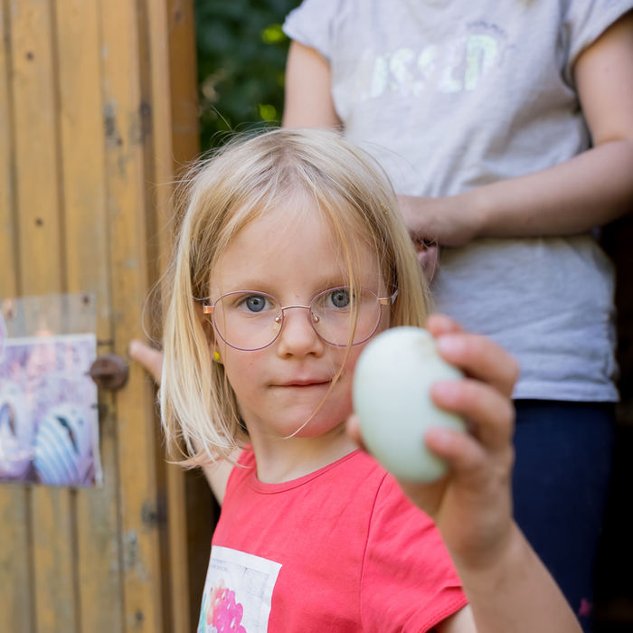 Blondes Kind hält Ei, zeigt Geschenkbox, trägt Brille, in Nähe einer Tür.