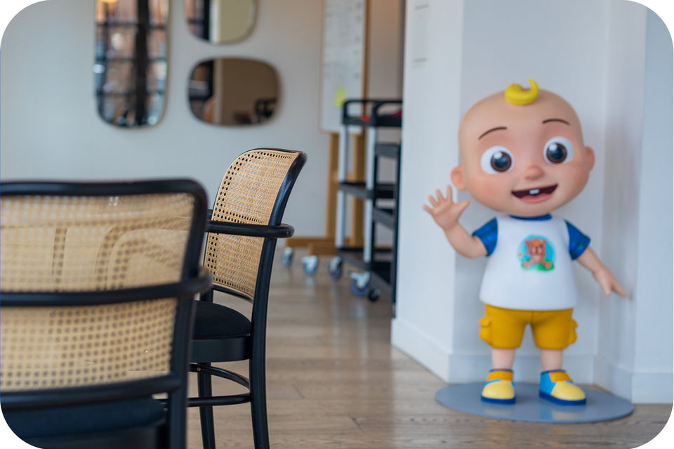 JJ, a CoComelon character, stands indoors, waving. 