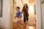 A toddler in a diaper runs towards the camera, while a woman holds a brown pants behind him.