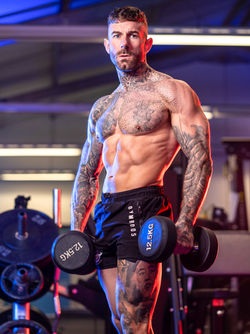 Muscular man holding dumbbells, gym shorts with tattoo arms, working out in the gym