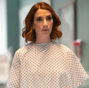 Mujer de cabello castaño en bata de hospital, de pie en un pasillo con fondo desenfocado. Expresión seria, ambiente clínico.