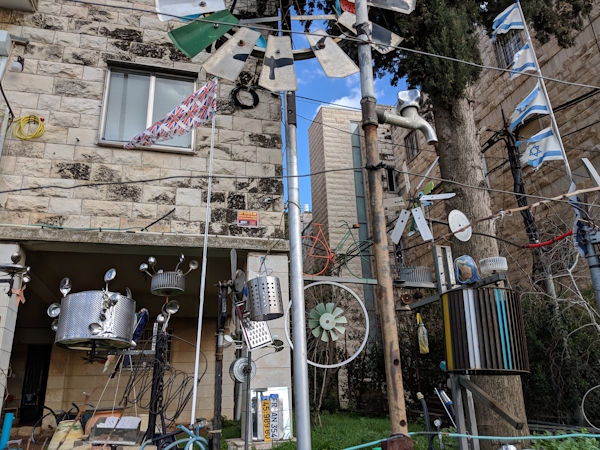 חצר של בית משותף בירושלים שיש בה פסלים קינטיים עשויים מכל מיני חומרים כמו כלי אוכל, תוף של מכונת כביסה, פומפייה ועוד. כולם זזים בכוח הרוח כמו סוג של שבשבות. צילום עדי ברטשניידר