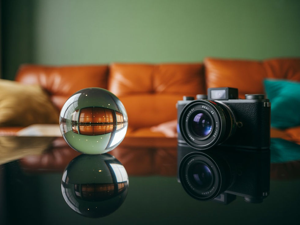 כדור זכוכית מונח על שולחן מבריק ולצידו מצלמה דיגיטלית שחורה עם עדשה גדולה, כששניהם משתקפים על פני השולחן. ברקע נראה סלון ביתי עם ספה חומה וקיר ירקרק, מטושטש בעומק שדה רדוד, והאור הטבעי יוצר תחושה של צילום, השתקפות ונקודת מבט משתנה. איור AI