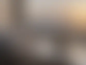 نافذة كبيرة تطل على شروق ذهبي فوق البحر، وبجانبها كوب شاي وقماش رمادي مطوي على الحافة في مشهد هادئ.