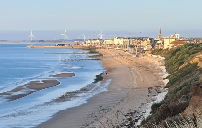 Promenade along Bridlington beach with Victorian architecture and distant pier under a partly cloudy sky.