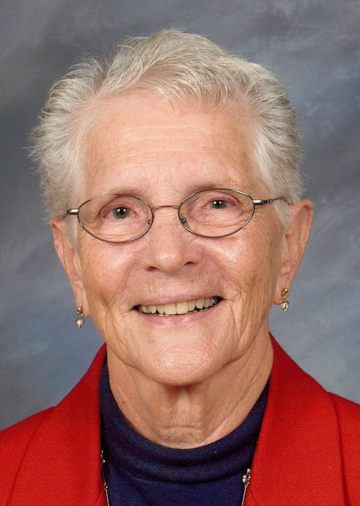 Lucille Ellen "Lucy" Hahn (née Krause) wearing glasses, red jacket, and dark top. Gray background. Appears cheerful and content.