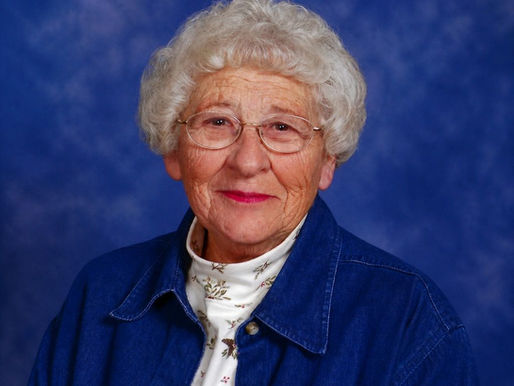Ardella D. Pethke Obituary photo with curly white hair, glasses, and a denim shirt smiles warmly against a blue mottled background.