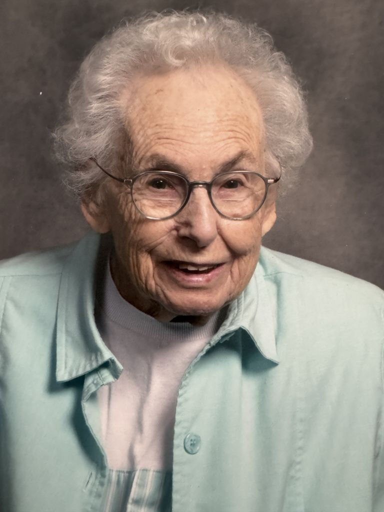 Joyce Joan Ponto Obituary photo with glasses and a light blue shirt, smiling against a gray background, conveying warmth and friendliness.