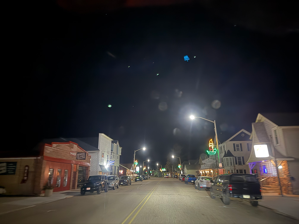 Nighttime street scene with lit storefronts, parked cars, and streetlights illuminating the road. A neon sign is visible in the distance.