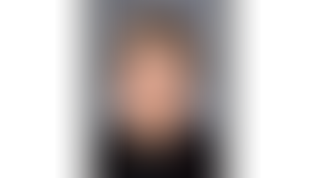 Eloyious I. Bonikowske with short curly hair, wearing a dark top, smiles softly against a gray background. Calm demeanor.