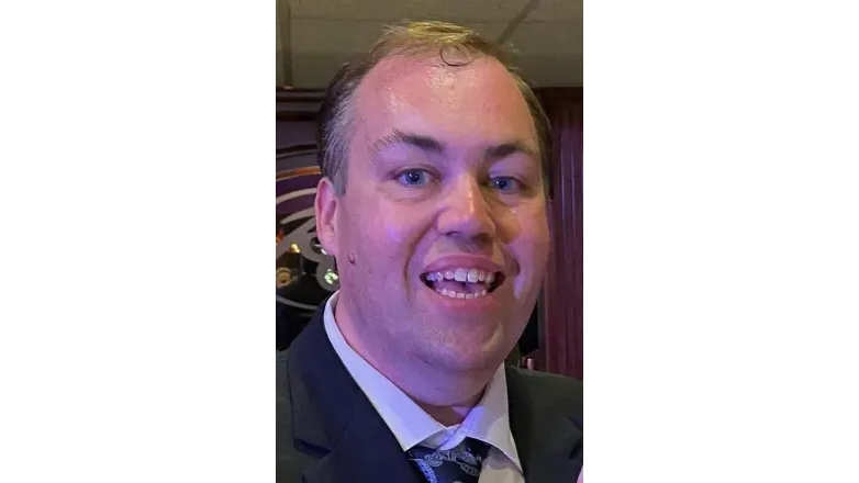Matthew Voight in a suit at an indoor event with purple lighting. Background shows blurred décor. jovial atmosphere.