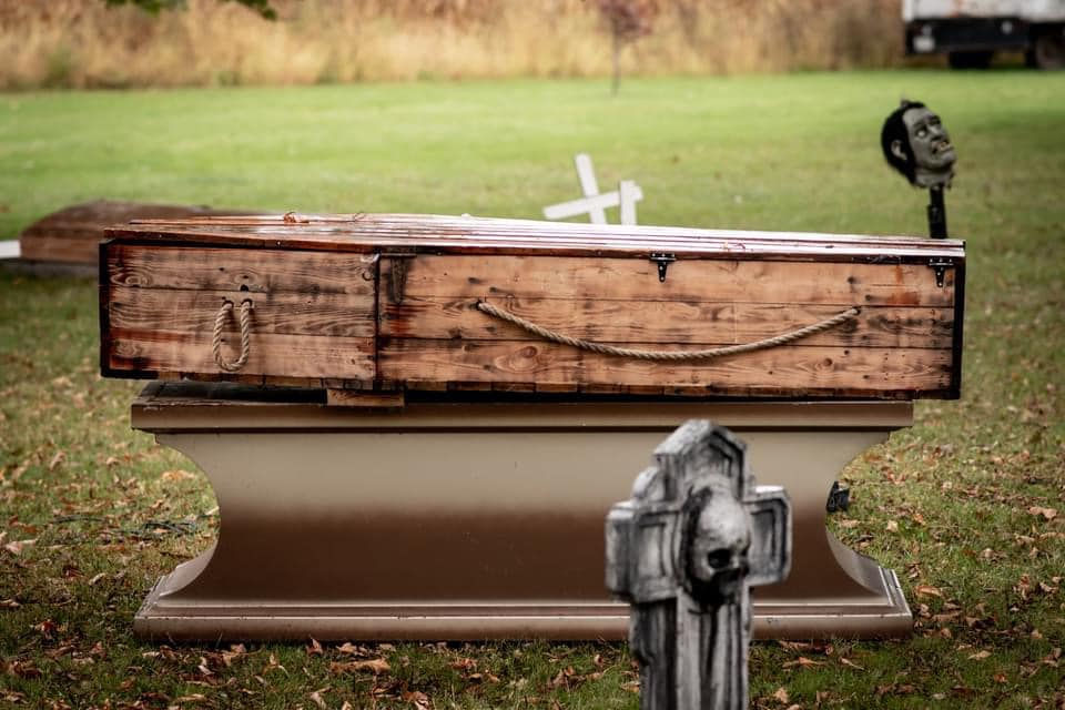 Wooden coffin on a beige platform in a grassy area, with a cross and skull decoration nearby, evoking a somber mood.