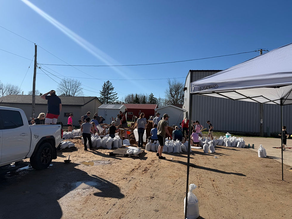 People filling sandbags under a blue sky near buildings. A white truck and canopy are visible. The mood is busy and communal.