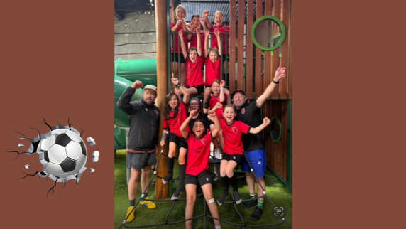 Image of a girls' soccer team wearing red uniforms, with their two coaches on either side wearing black. They're on a playground with their arms in the air, cheering.