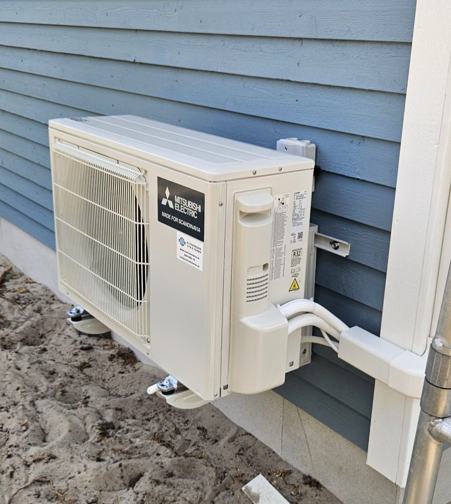 Mitsubishi Electric värmepump installerad vid blå träfasad i Kristianstad. Modern luft/vatten-värmepump som ger låg energiförbrukning och hög komfort året runt.