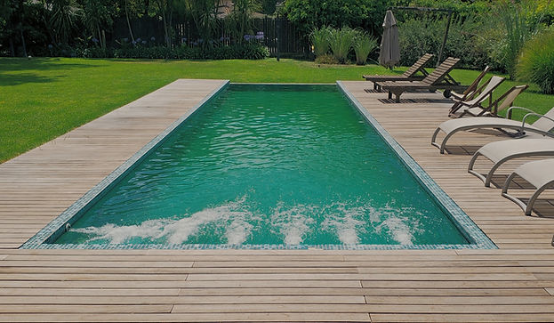 Piscina rectangular premium con borde de madera y sistema de hidromasaje, rodeada de reposeras en un parque arbolado.