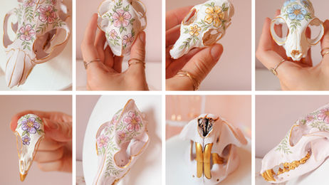 Hand holding a collage of painted animal skulls, white with pink, blue, and yellow floral designs and gold accents on a soft pink background