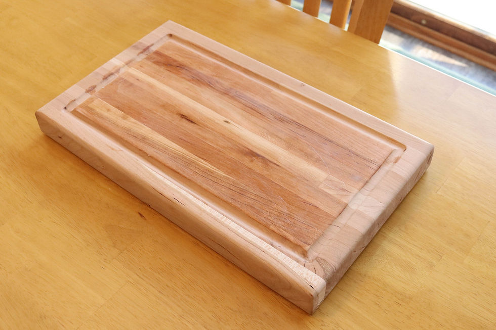 Wooden cutting board on a light wooden table. The board has a smooth, polished surface with visible grain patterns, creating a warm, rustic feel.