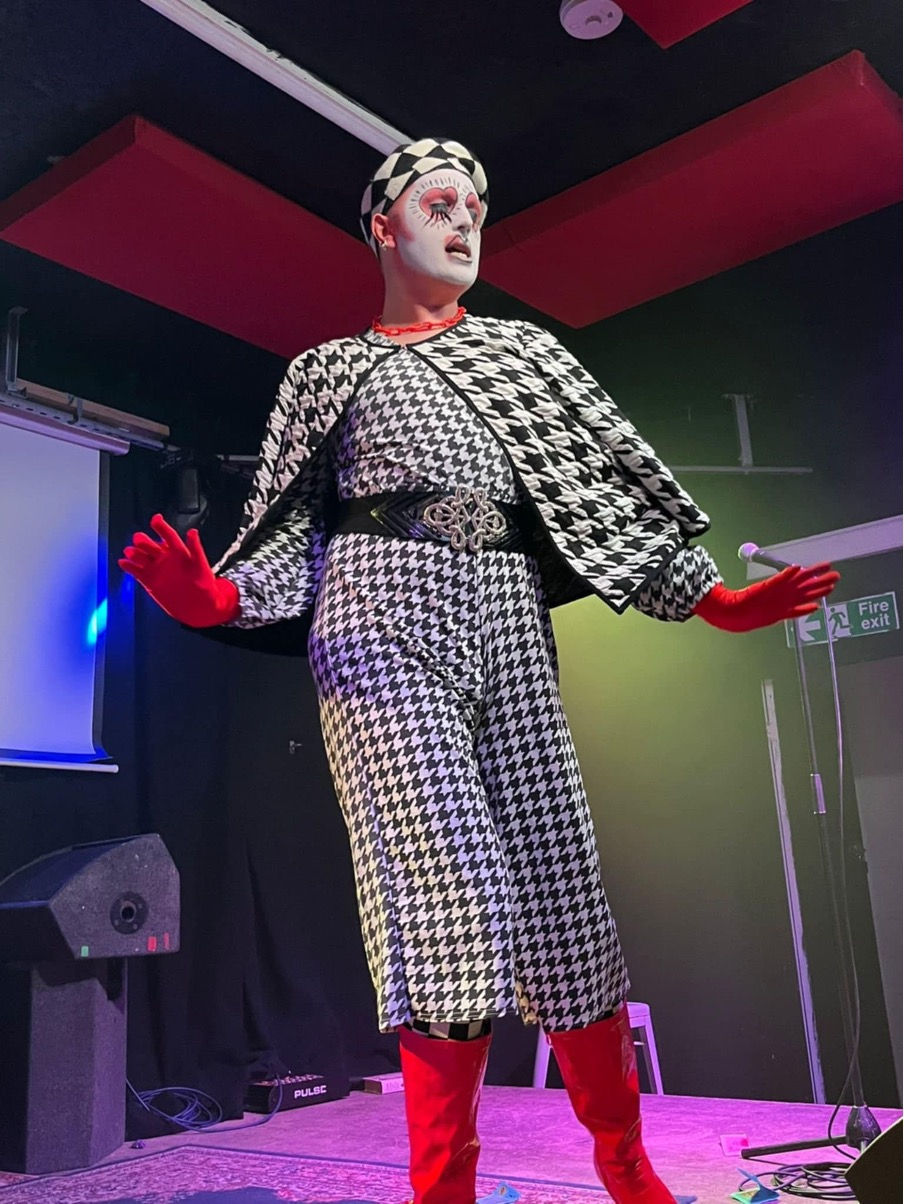 Pop Tart lip syncs on stage. She's tall, in a monochrome houndstooth dress suit and cape, red gloves and boots, monochrome beret, and white ace paint with love heart eyes, like a pop art work. It's striking and stunning.