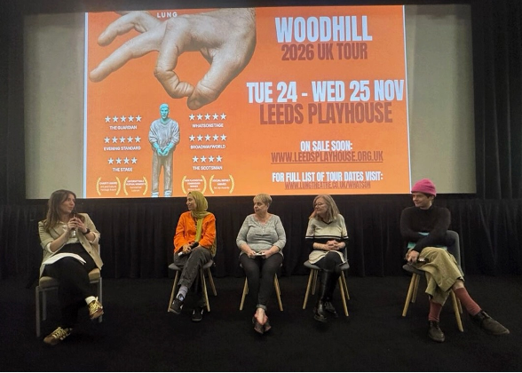 Four panel members and a host seated on stage in front of a cinema screen displaying the Woodhill film advertisement. The panel is taking part in a discussion about deaths in custody, accountability, law and institutional practice.