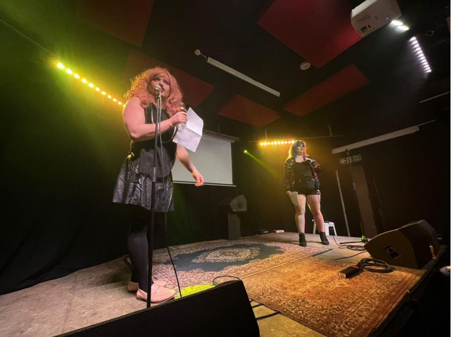 Alice Aforethought and Fox Populi narrating the legal history onstage. Alice in pink long wig, dark top and shirt, pink shoes, holding a script while she speaks to the audience. Fox is in a black romper and rainbow sequin jacket, pink and blue hair, by another microphone, also engaging with the audience. They are conveying the history of queerness and law between acts.