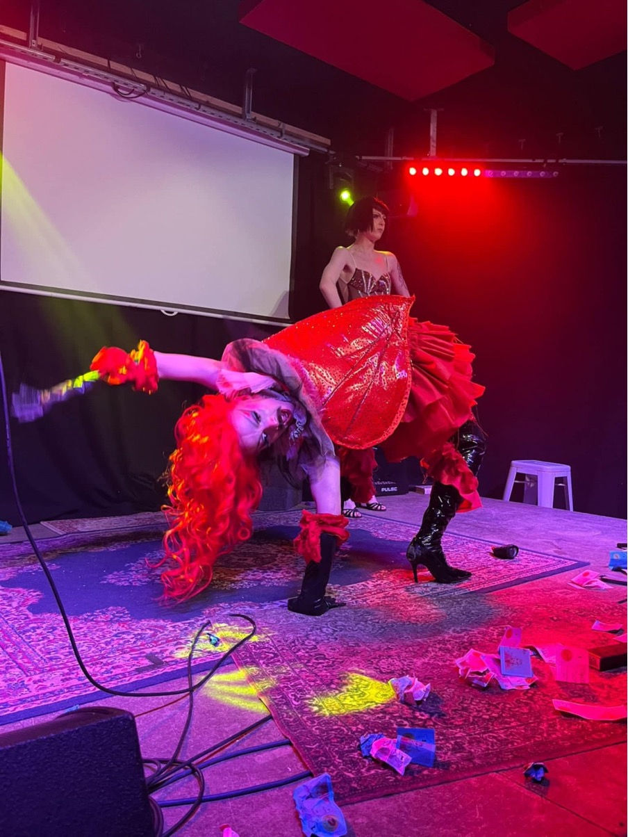 Pembo doing stunts onstage. Red wig, red valentine's heart corset and red dress, knee length black boots and matching gloves, dark moustache on feminine make up. She is arched backwards, seemingly with one hand and one boot on the floor, mid-manoeuvre. House down boots, as they say.