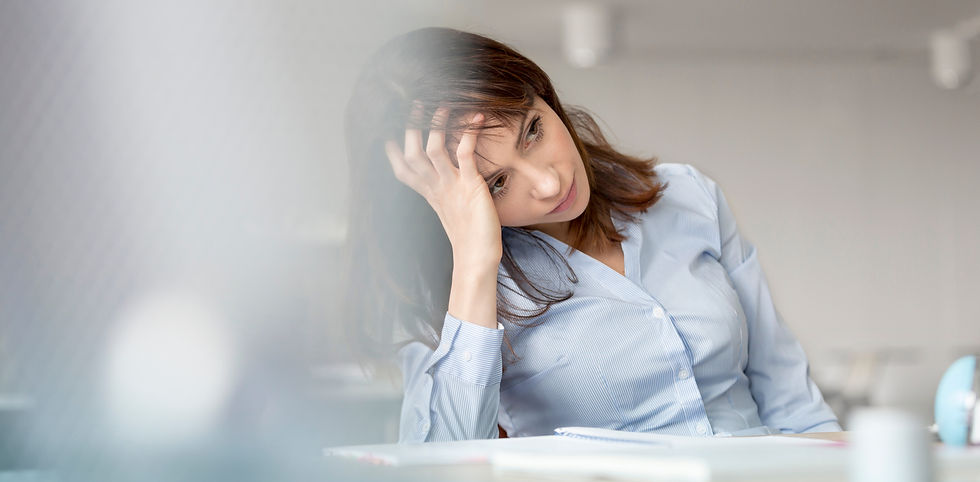 Woman in a blue blouse sits at a desk, looking stressed with her head resting on hand. Blurred office background enhances mood.