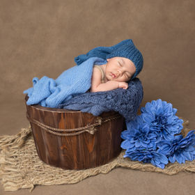 Newborn baby boy sleeping in a rustic wooden bucket with soft blue knit wrap, matching hat, and blue flowers, photographed during a San Diego newborn photoshoot.