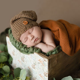 Newborn boy sleeping in wooden drawer wrapped in rust-orange fabric, wearing brown knitted hat, with green leaves nearby, photographed in San Diego baby photography studio.