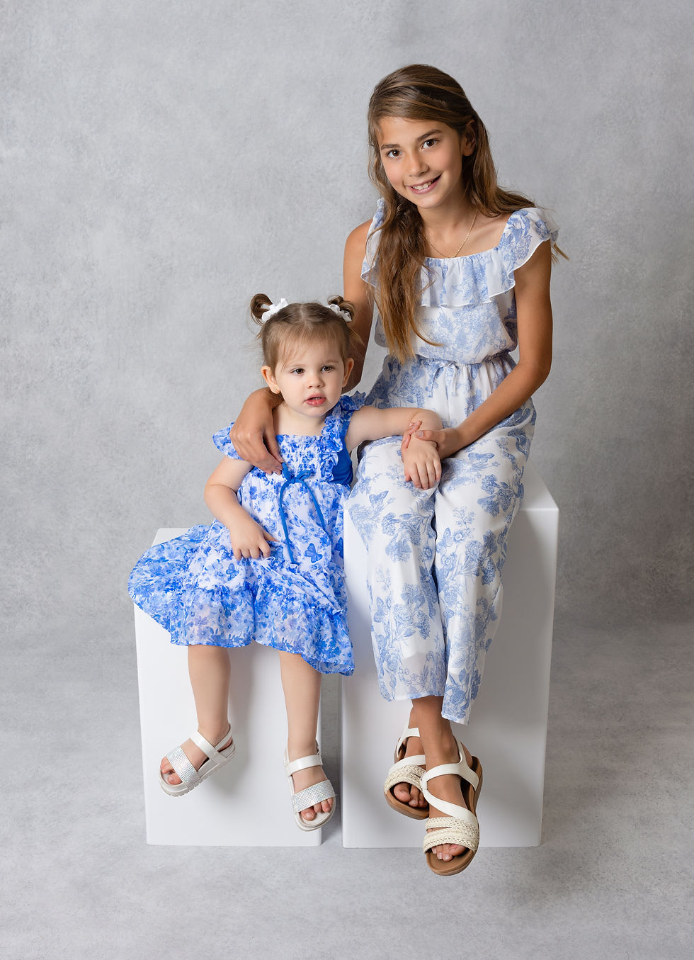 Toddler girl in blue floral dress sitting on a white cube with her older sister in matching outfits for a birthday session in San Diego photography studio