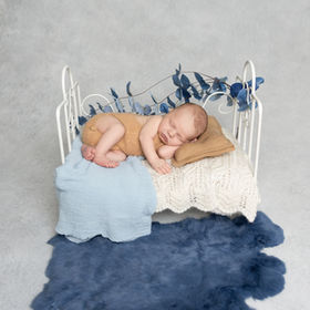 Newborn baby boy in beige romper sleeping on white metal bed with blue accents, captured in San Diego newborn photography studio.