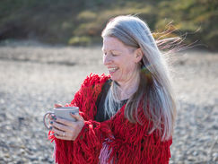 Somatic Therapist Laura Starky wearing a red fringed jacket holding a mug, smiling on a sunny beach with wind in her hair. Pebbly shore and greenery in the background, healing practice for embodied women.