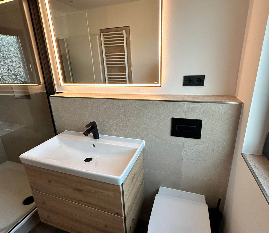 Kompaktes Badezimmer mit Waschtisch in Holzoptik, wandhängendem WC, LED-Spiegel und schwarzer Drückerplatte auf heller Wandverkleidung.