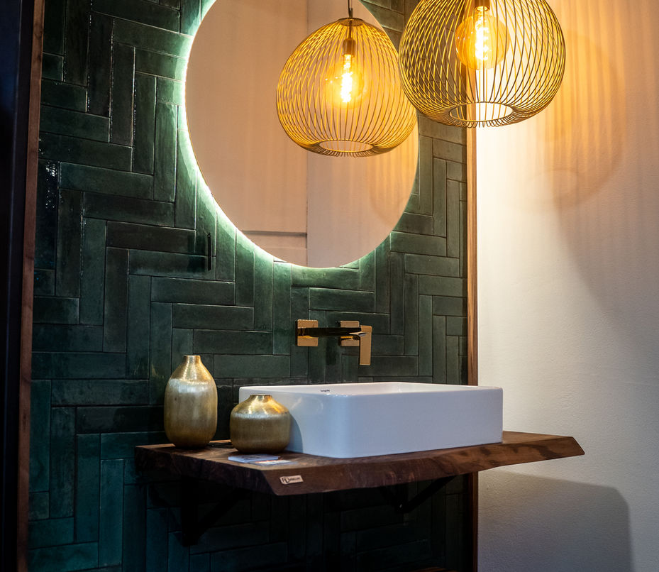 Design-Waschplatz mit grünem Fliesenpanel im Fischgrätmuster, schwebendem Naturholz-Waschtisch, Rundspiegel mit LED-Licht und goldenen Hängelampen.