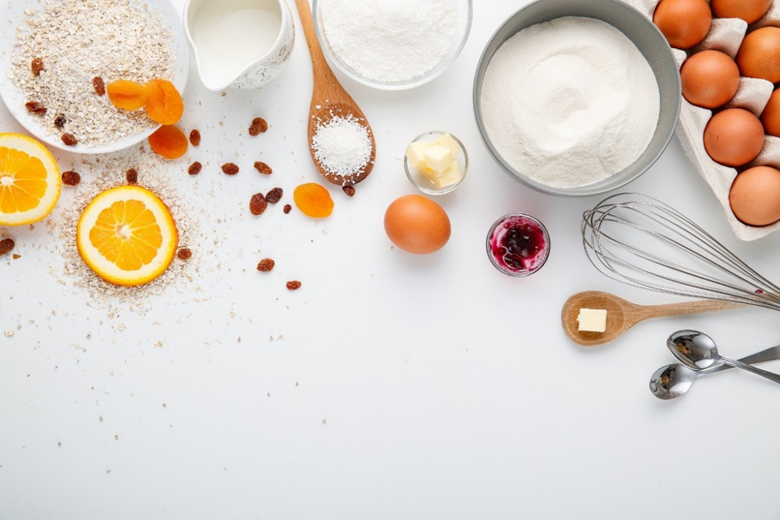 A flatlay of baking ingredients including flour, sugar, eggs, butter, and oranges.
